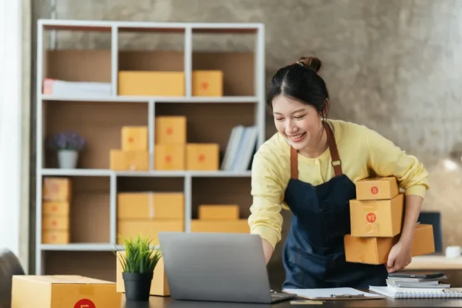 An Asia Woman Entrepreneur Carrying Boxes While Working on a Laptop
